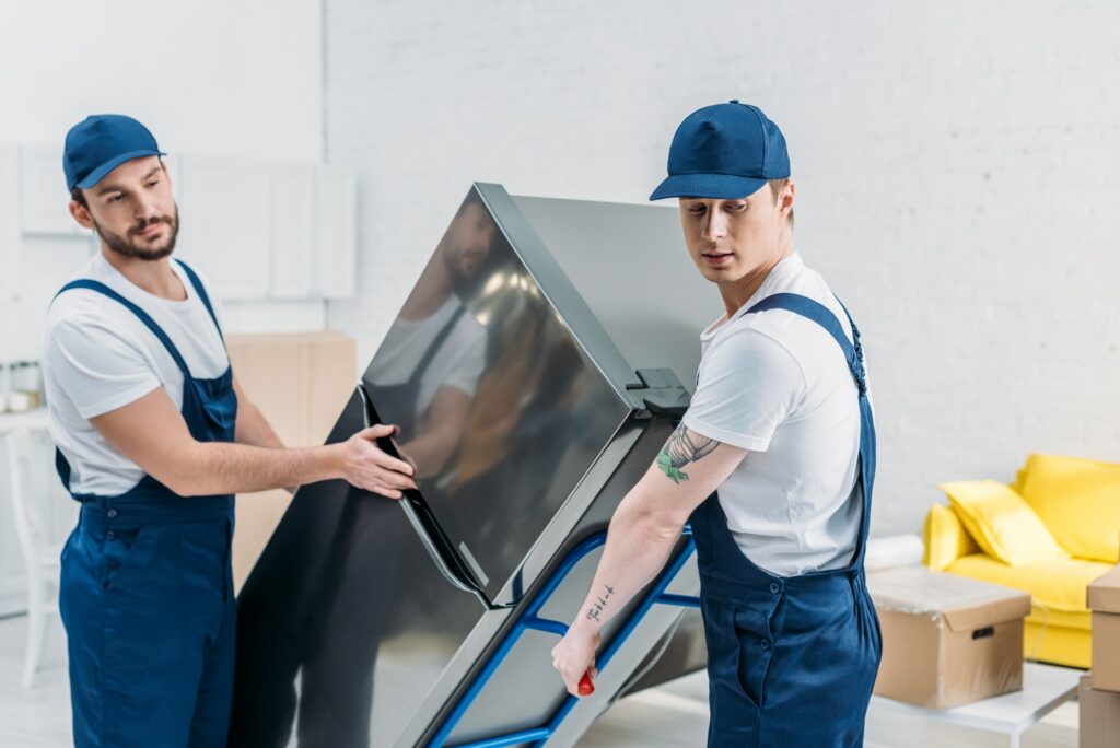First Class Movers LLC. two-movers-in-uniform-using-hand-truck-while-transporting-refrigerator-in-apartment